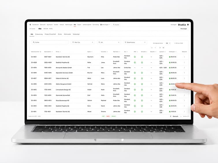 Studio-M-Backoffice auf dem Laptop: Abo-Liste mit Abonummer, Firma, Abo-Typ und Laufzeit, eine Hand zeigt auf einen Eintrag